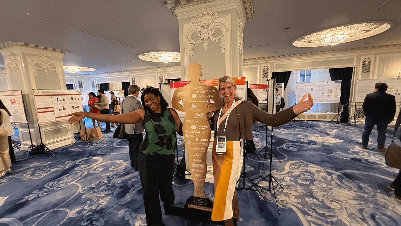 Two women posing happily beside a large bottle sculpture indoors.